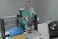 Cycles render of the tool head on the 3018: teal body, white printed gears, the syringe with a yellow tip, the USB microscope and the orange probe arm over the beaker matrix